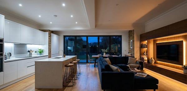 Evening photo of a UK open plan room with distinct lighting zones: cool white in the kitchen and warm amber from media wall LEDs in the living area.